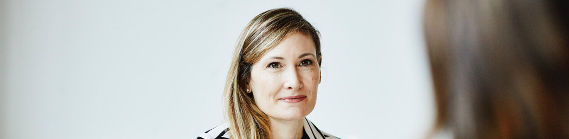Eine Frau mit langen blonden Haaren unterhält sich an ihrem Schreibtisch mit einer Patientin.