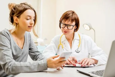 Eine Ärztin mit weißem Kittel und gelbem Stethoskop um den Hals sitzt neben einer Patientin an einem Tisch. Die Patientin hält ein Smartphone in der Hand, beide schauen auf den Bildschirm.