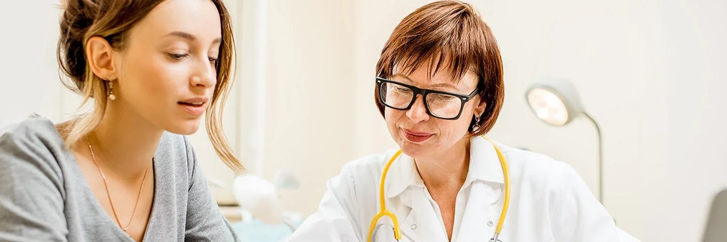 Eine Ärztin mit weißem Kittel und gelbem Stethoskop um den Hals sitzt neben einer Patientin an einem Tisch. Die Patientin hält ein Smartphone in der Hand, beide schauen auf den Bildschirm.