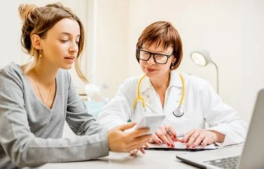 Eine Ärztin mit weißem Kittel und gelbem Stethoskop um den Hals sitzt neben einer Patientin an einem Tisch. Die Patientin hält ein Smartphone in der Hand, beide schauen auf den Bildschirm.