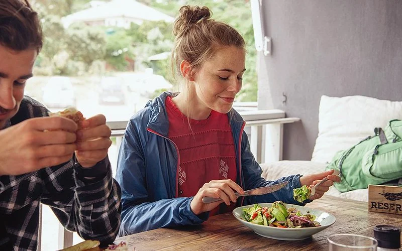 Ein Mann und eine Frau sitzen nebeneinander an einem rustikalen Holztisch. Der Mann isst ein Stück helles Brot, die Frau isst buntes Gemüse aus einer grauen Keramikschale mit Messer und Gabel.