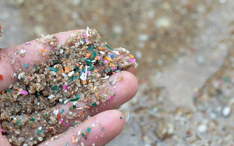 Hand mit nassem Sand, in dem viele kleine Plastikpartikel sichtbar sind.