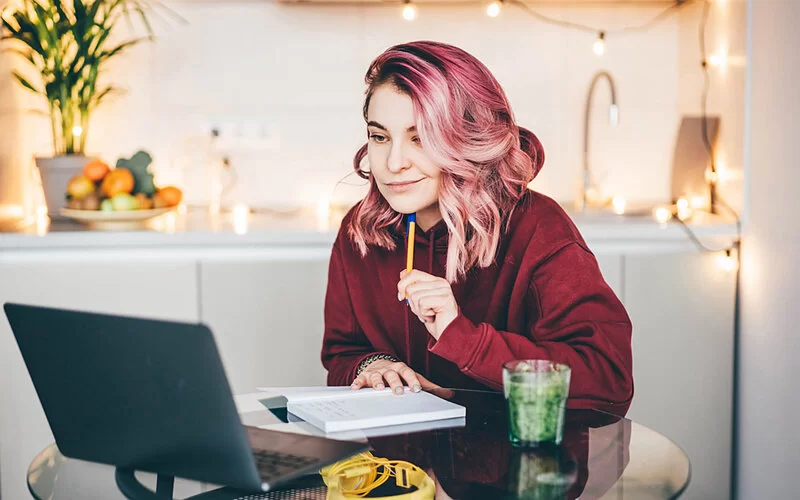 Eine junge Frau sitzt an einem Tisch vor ihrem Laptop und macht sich Notizen in einem kleinen Schreibheft.