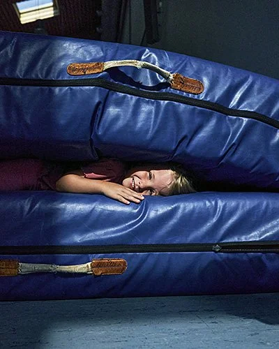 Ein junges Kind lächelt fröhlich zwischen zwei großen, übereinander gestapelten, blauen Turnmatten hervor. Die Matten haben braune Lederriemen als Griffe. Der Hintergrund ist dunkel.