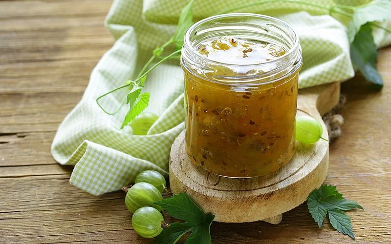 Ein offenenes Schraubglas mit selbstgemachter Stachelbeermarmelade auf einem runden Holzbrettchen auf einem rustikalen Holztisch, der mit einem hellgrün-weiß-karierten Küchentuch, einigen ganzen Stachelbeeren und Stachelbeergrün dekoriert ist.
