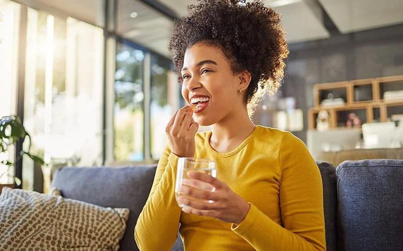 Eine Frau in einem gelben Oberteil hält ein Glas Wasser in der Hand und nimmt eine Kapsel ein.