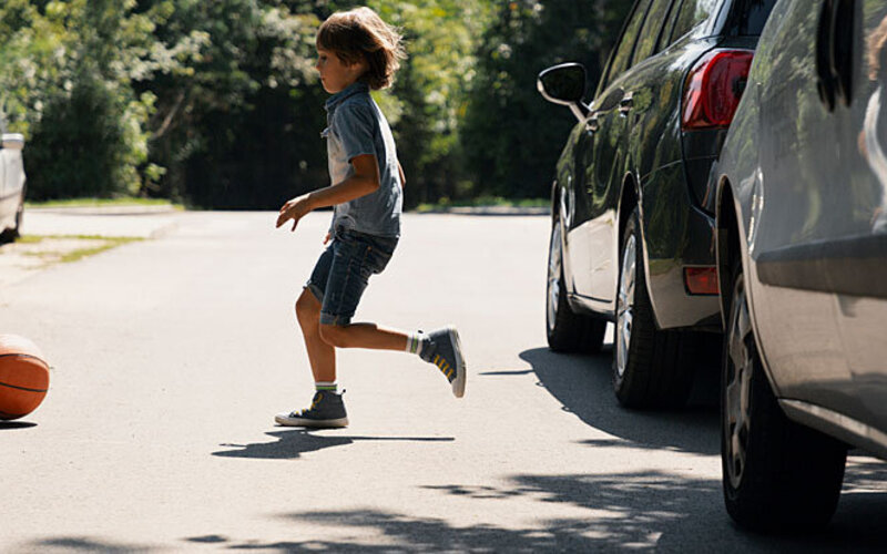 Unbeschwerter Junge läuft hinter dem Ball auf der Straße neben Autos.