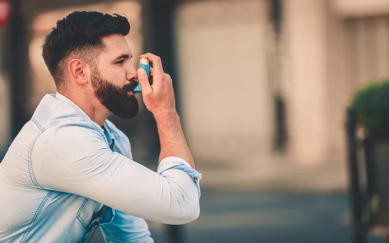 Ein junger Mann mit Vollbart hält sich ein Asthma-Inhalator an den Mund.