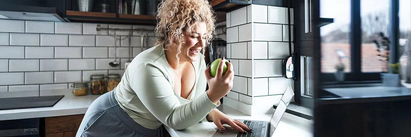 Eine junge Frau steht an einer Küchentheke, schaut auf den Bildschirm eines Laptops und issst dabei einen Apfel.