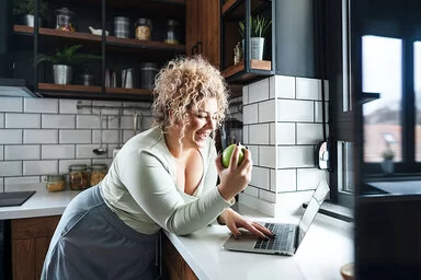 Eine junge Frau steht an einer Küchentheke, schaut auf den Bildschirm eines Laptops und issst dabei einen Apfel.