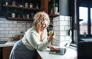 Eine junge Frau steht an einer Küchentheke, schaut auf den Bildschirm eines Laptops und issst dabei einen Apfel.