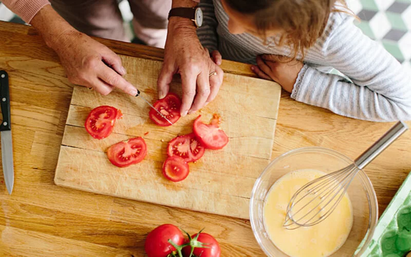 Großmutter und Enkelkind bereiten zusammen Abendbrot vor, sie schneiden Tomaten auf einem Brett.