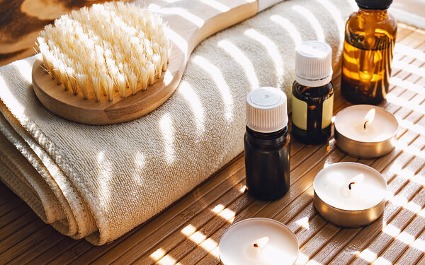 Auf einem zusammengerollten Handtuch liegt eine Massagebürste mit Holzgriff, daneben stehen diverse ätherische Öle und eine Kerze.