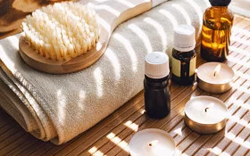Auf einem zusammengerollten Handtuch liegt eine Massagebürste mit Holzgriff, daneben stehen diverse ätherische Öle und eine Kerze.