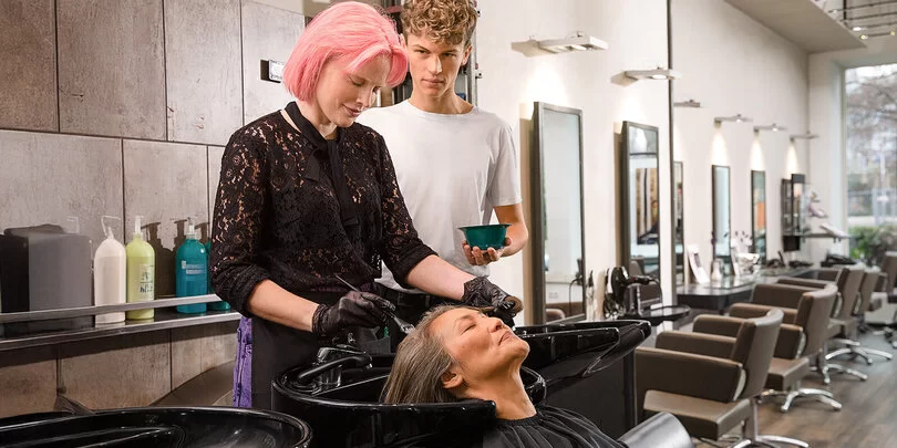 Eine Frau mit pinken Haaren färbt einer Kundin im Friseursalon die Haare. Die Kundin sitzt entspannt an einem schwarzen Waschbecken. Ein junger Mann steht daneben und hält eine Schale mit Haarfarbe. Im Hintergrund sind weitere Friseurstühle und Spiegel in einem modern eingerichteten Salon zu sehen.