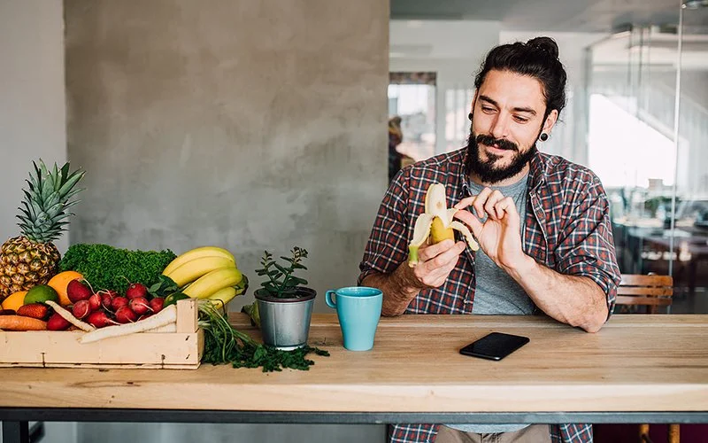 Ein junger Mann mit Zopf sitzt an einer Theke und schält eine Banane. Auf der Theke steht ein Korb mit Obst und Gemüse.