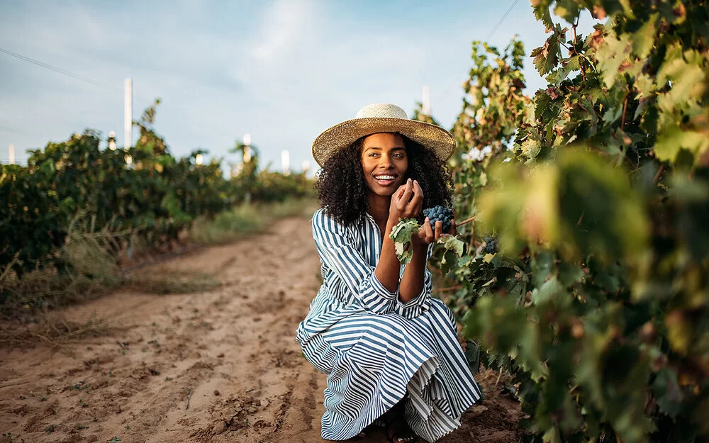 Junge Frau in einem Weinberg geht vor einer Weinrebe in die Hocke und probiert die roten Trauben.