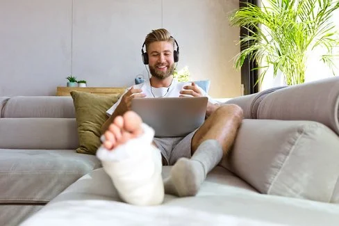 Lächelnder junger Mann mit gebrochenem Bein in Gipsverband liegt zu Hause auf dem Sofa aund schaut auf den Bildschirm seines Laptops.