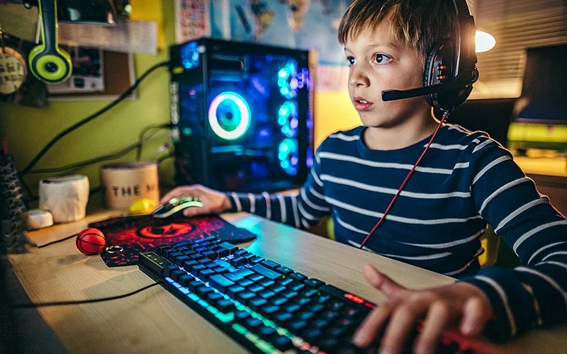 Ein Junge sitzt an einem Tisch vor einer Computertastatur. Er bedient die Maus mit der rechten Hand und die Tastatur mit der linken. Er trägt ein Headset und blickt auf den Monitor des PCs, der nicht abgebildet ist, aber sein Gesicht beleuchtet. Neben dem Tisch steht ein blau leuchtender Gaming-PC.