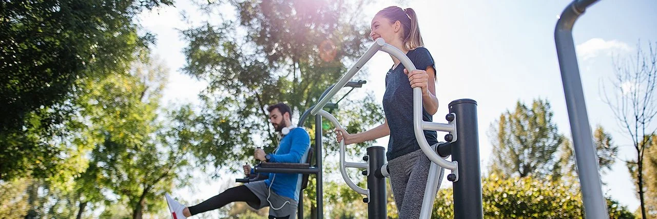 Ein junger Mann und eine junge Frau in Sportkleidung trainieren in einem Park an Fitnessgeräten.