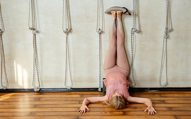 Eine Frau in sportlicher Kleidung liegt mit den Füßen nach oben vor einer Wand und hat Sandsäcke auf den Füßen.
