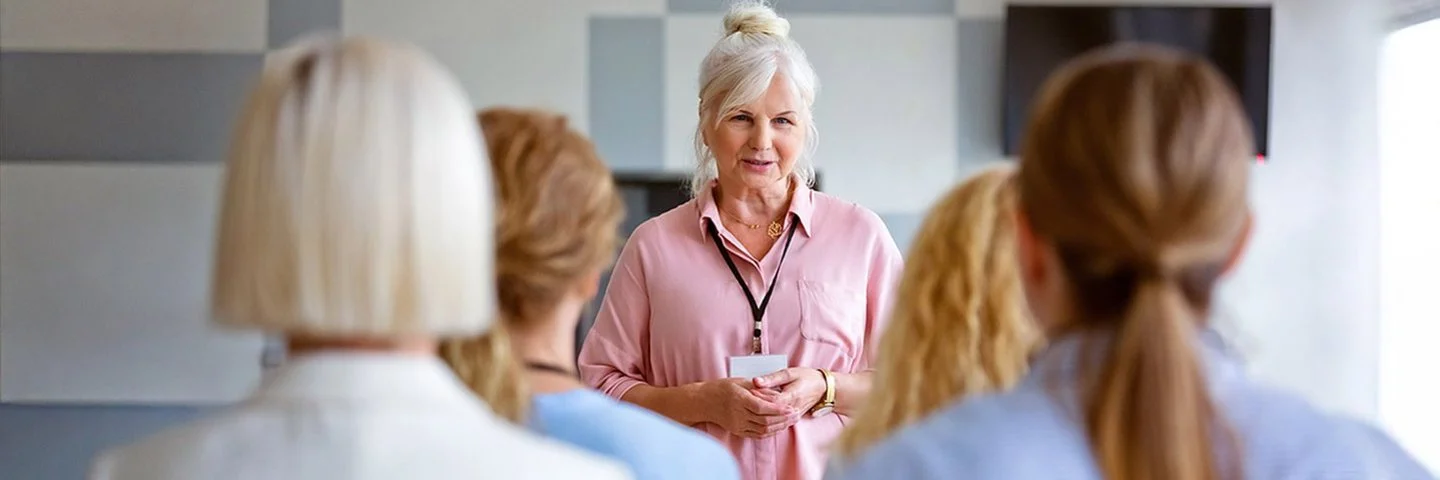 Eine ältere Frau, steht vor einer Gruppe und gibt eine Schulung.