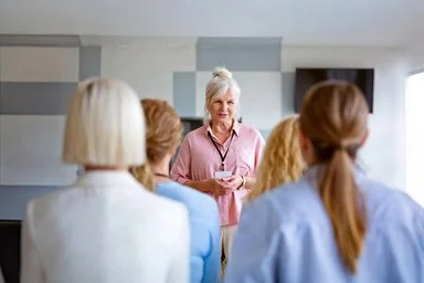 Eine ältere Frau, steht vor einer Gruppe und gibt eine Schulung.