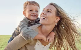 Eine Mutter trägt ihren kleinen Sohn huckepack über eine Wiese und beide lachen dabei.