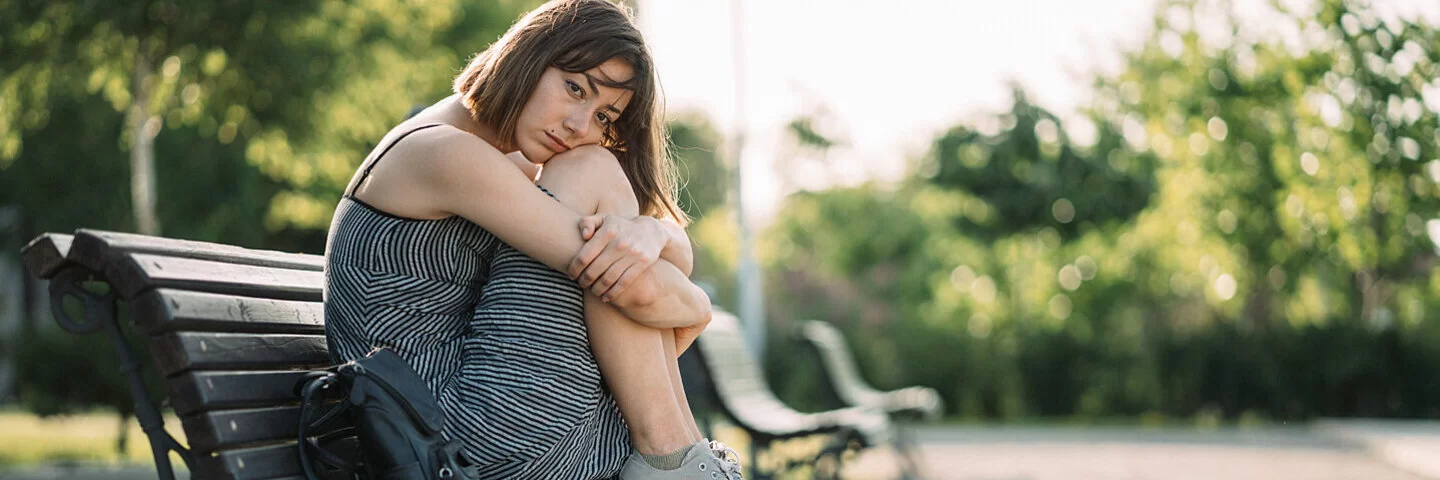 Eine junge Frau sitzt mit traurigem Gesichtsausdruck im Sommer auf einer Parkbank.