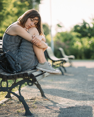 Eine junge Frau sitzt mit traurigem Gesichtsausdruck im Sommer auf einer Parkbank.