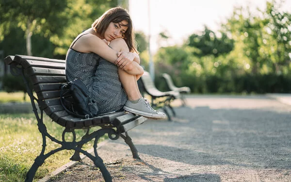 Eine junge Frau sitzt mit traurigem Gesichtsausdruck im Sommer auf einer Parkbank.