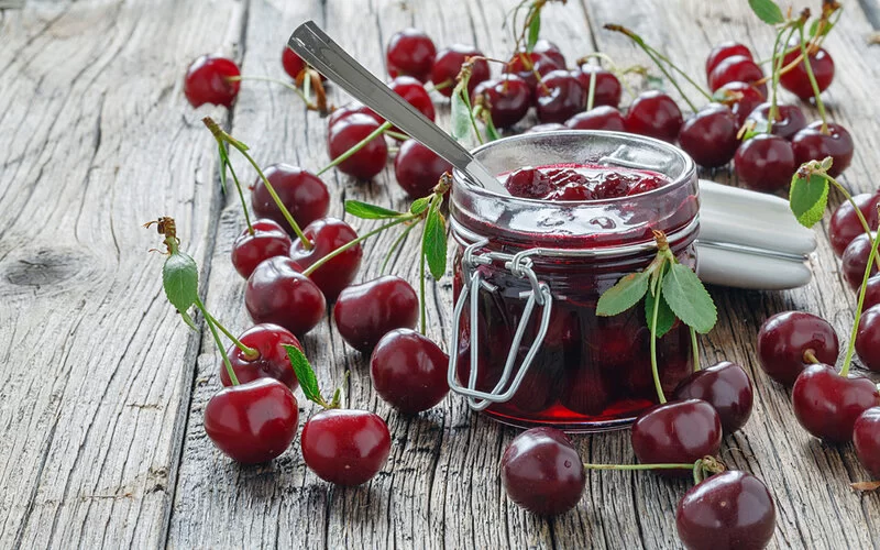 Ein Glas Kirschmarmelade steht auf einem hellen Holztisch, rundherum liegen frische Kirschen.