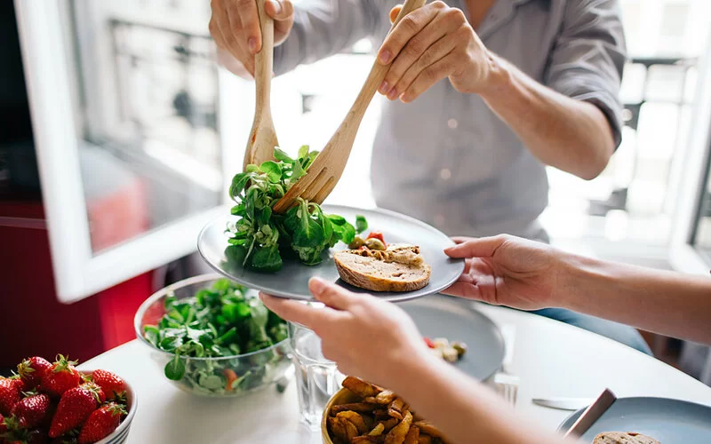 Gedeckter Tisch, mit Salat und Obst. Eine Person gibt der anderen Salat auf einen Teller mit Brot und Oliven auf.