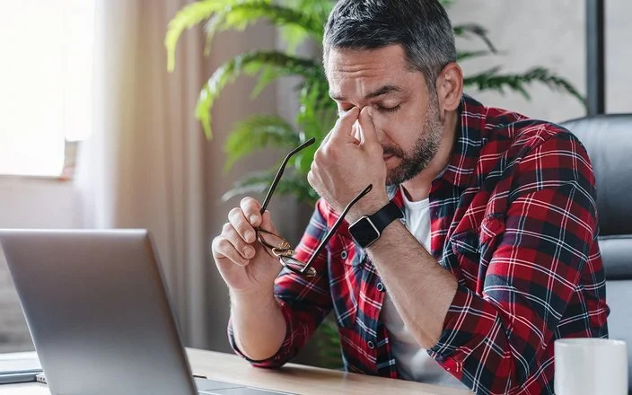 Ein gestresst wirkender Mann sitzt vor einem Laptop, er hält in der einen Hand seine Brille und fasst sich mit der anderen Hand an das Nasenbein.