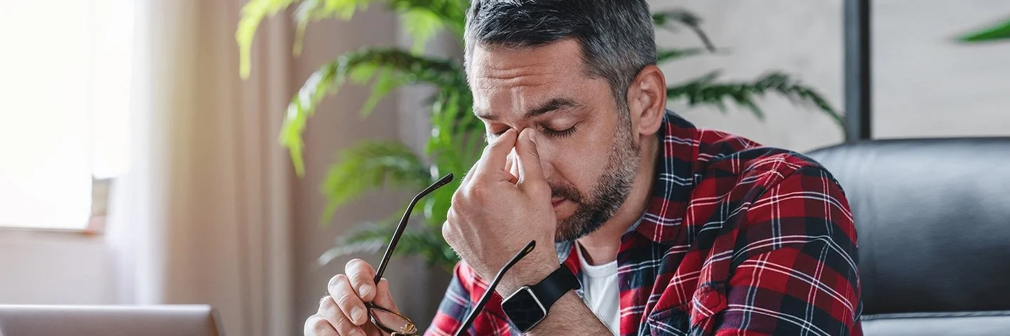 Ein gestresst wirkender Mann sitzt vor einem Laptop, er hält in der einen Hand seine Brille und fasst sich mit der anderen Hand an das Nasenbein.