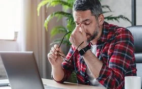 Ein gestresst wirkender Mann sitzt vor einem Laptop, er hält in der einen Hand seine Brille und fasst sich mit der anderen Hand an das Nasenbein.