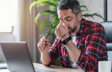 Ein gestresst wirkender Mann sitzt vor einem Laptop, er hält in der einen Hand seine Brille und fasst sich mit der anderen Hand an das Nasenbein.