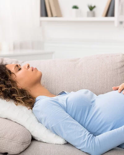 Eine junge schwangere Frau liegt auf der Couch und hält sich den schmerzenden Bauch.