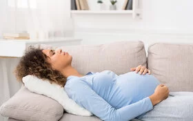 Eine junge schwangere Frau liegt auf der Couch und hält sich den schmerzenden Bauch.