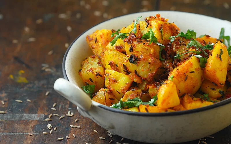 Auf einem Holztisch steht eine Schale mit dem traditionellen indischen Kartoffelgericht Jeera Aloo. Die angebratenen Kartoffeln sind mit Kräutern und Kreuzkümmelsamen bestreut. Weitere Samen liegen verstreut um die Schüssel herum auf dem Tisch.
