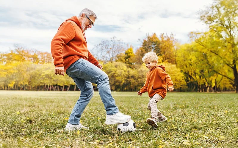 Ein ca. 55-jähriger Mann mit Vollbart und Brille spielt mit einem ca. 4-jährigen blonden Jungen Fußball in einem Park.