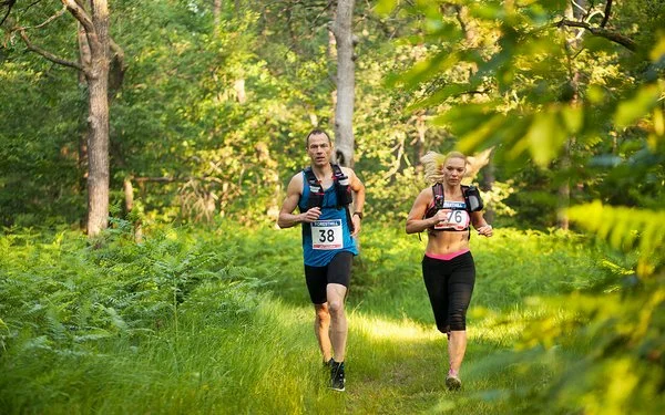 Ein Mann und eine Frau in kurzer Sportkleidung und umgeschnallten Trinkflaschen laufen durch einen Wald.