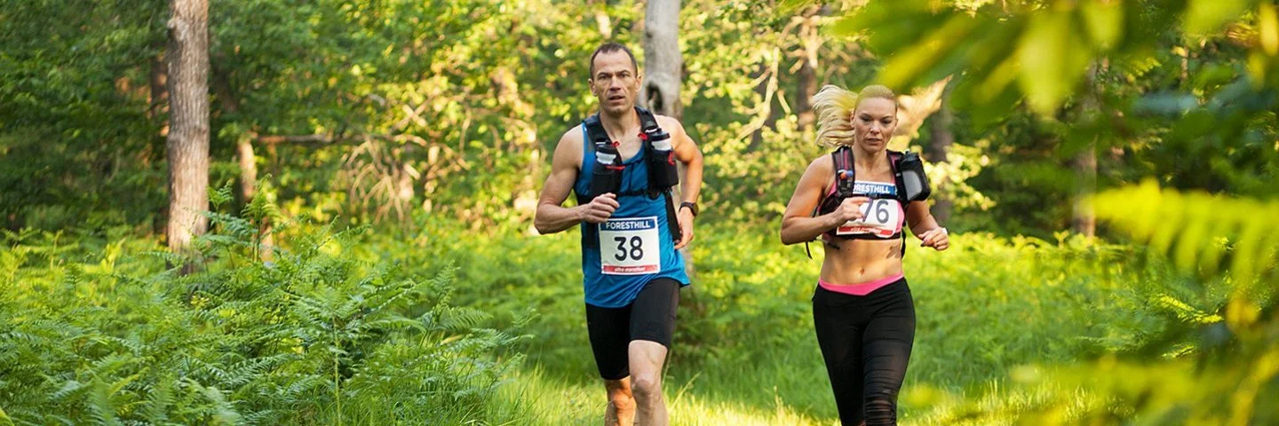 Ein Mann und eine Frau in kurzer Sportkleidung und umgeschnallten Trinkflaschen laufen durch einen Wald.