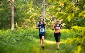 Ein Mann und eine Frau in kurzer Sportkleidung und umgeschnallten Trinkflaschen laufen durch einen Wald.