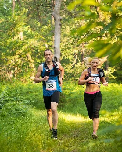 Ein Mann und eine Frau in kurzer Sportkleidung und umgeschnallten Trinkflaschen laufen durch einen Wald.