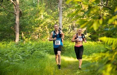 Ein Mann und eine Frau in kurzer Sportkleidung und umgeschnallten Trinkflaschen laufen durch einen Wald.