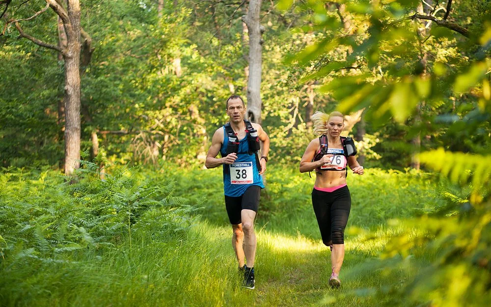 Ein Mann und eine Frau in kurzer Sportkleidung und umgeschnallten Trinkflaschen laufen durch einen Wald.