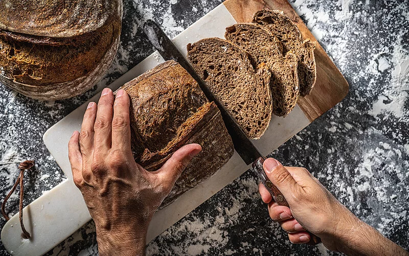 Ein nicht erkenntlicher Mann schneidet Vollkornbrot auf einem Brett in Scheiben.