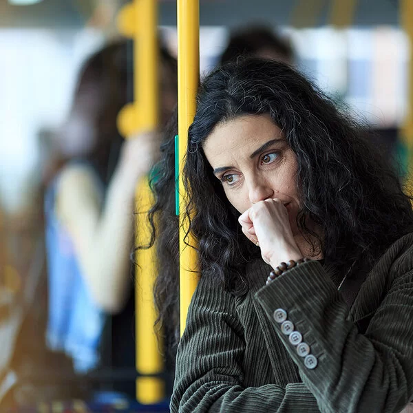 Eine junge Frau steht in einem Bus. Sie lehnt sich an eine gelbe Haltestange, dabei scheint sie intensiv zu grübeln.