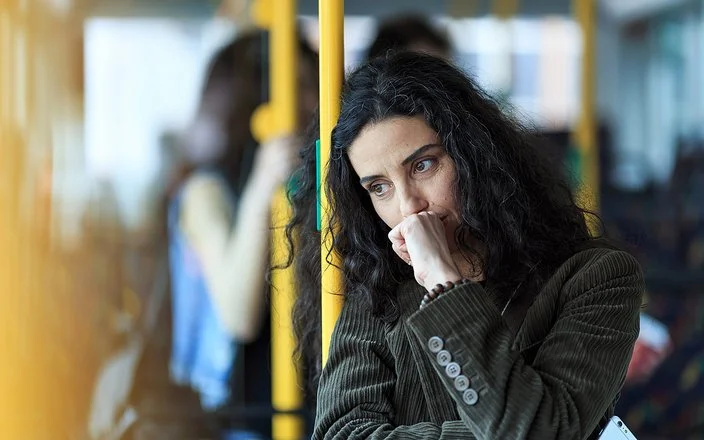 Eine junge Frau steht in einem Bus. Sie lehnt sich an eine gelbe Haltestange, dabei scheint sie intensiv zu grübeln.